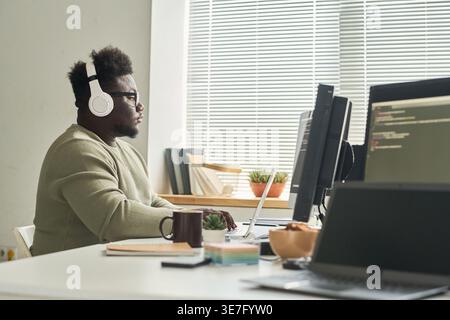 Serious Black male programmer wearing wireless headphones using laptop while sitting at table with coffee in office Stock Photo