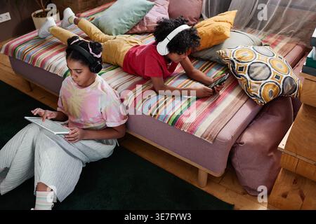 Black boy with curly afro wearing headphones using smartphone lying down on bed near his sister using tablet sitting on floor in bedroom at home Stock Photo