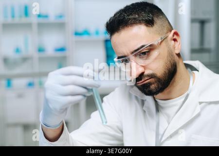 Middle Eastern man wearing white lab coat and gloves checking test tube with blue substance sitting in laboratory Stock Photo