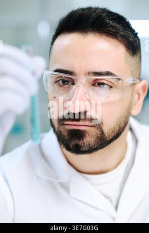 Middle Eastern man wearing white lab coat and gloves checking test tube with blue substance in laboratory Stock Photo