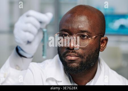 Black bald man wearing lab coat and gloves checking test tube with blue liquid substance sitting in laboratory Stock Photo