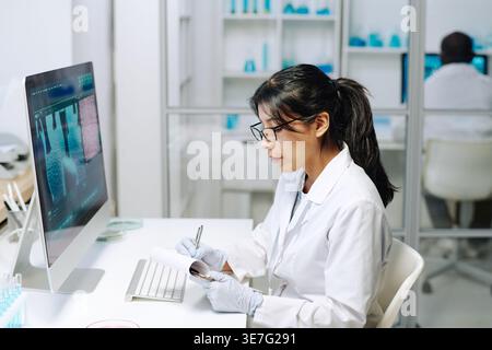 Hispanic woman wearing white lab coat and eyeglasses taking notes on notepad sitting in front of desktop in laboratory Stock Photo