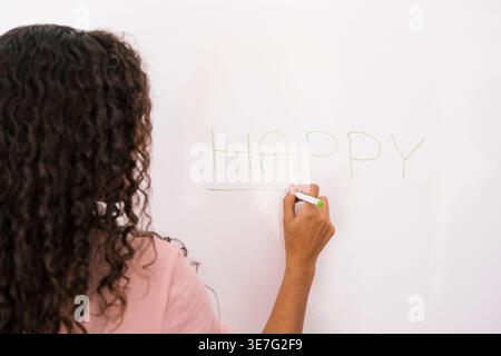 Hispanic female teacher with curly hair writing the word happy on whiteboard standing in classroom Stock Photo