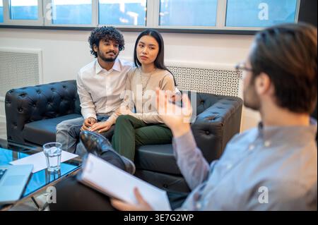 Smiling Indian male hugging Asian girlfriend sitting together on sofa attending therapy session with caucasian male psychotherapist sitting in clinic Stock Photo