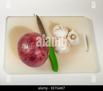 A red onion and three cloves of garlic on a cutting board with a green handled paring knife! Stock Photo