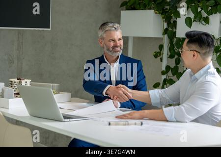 Asian male architect in eyeglasses shaking hands with caucasian male colleague after successful meeting while sitting at table with laptop and arch... Stock Photo