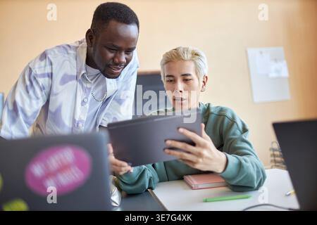 Asian male student sitting at table explaining data on tablet to Black male classmate standing beside him in classroom Stock Photo