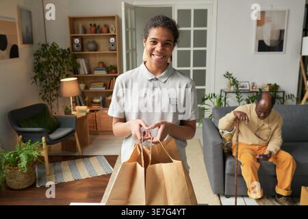 Black young female social service worker wearing uniform holding grocery bags standing near middle-aged Black man with cane using smartphone sittin... Stock Photo