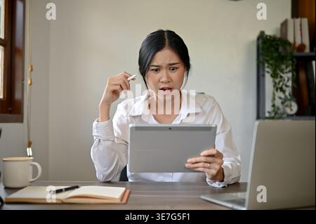 Shocked Asian female holding stylus pen and using tablet while sitting at table at home Stock Photo