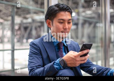 Smiling Asian businessman in blue suit using smartphone while sitting on floor at train station during daytime Stock Photo