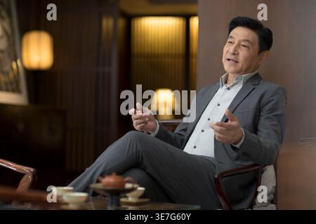 Cheerful Chinese businessman wearing gray suit sitting with legs crossed on chair in office Stock Photo