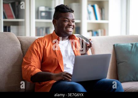 Smiling Black man with braids recording voice messages while using laptop sitting on sofa in living room at home Stock Photo