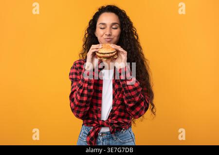 Smiling Middle Eastern woman with curly hair holding and smelling burger while standing with closed eyes against yellow background in studio Stock Photo