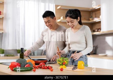 Asian woman mixing vegetable salad on bowl while standing at kitchen counter with her husband cooking at home Stock Photo