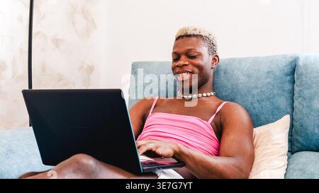 Smiling Black non-binary person using laptop sitting on sofa in living room at home Stock Photo
