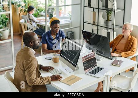Serious Black male programmer in eyeglasses sitting at desk with caucasian female colleague developing new software on computers working together i... Stock Photo