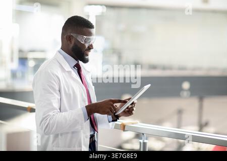 Black male medical scientist in labcoat and protective glasses using tablet standing in laboratory Stock Photo