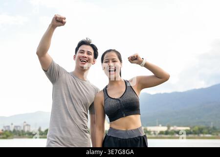 Cheerful Thai couple with raised arms and closed fists celebrating success after reaching their running target while standing together near lake in... Stock Photo