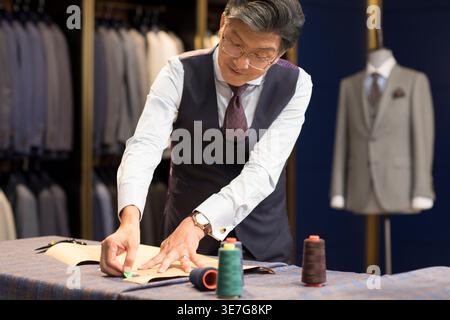 Senior Chinese male fashion designer standing at table working in his formal suits shop Stock Photo