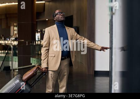 Black businessman with luggage wearing eyeglasses looking up pressing button for elevator while standing on hallway in hotel Stock Photo