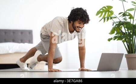 Asian man with curly hair watching fitness video on laptop while planking on yoga mat in bedroom at home Stock Photo