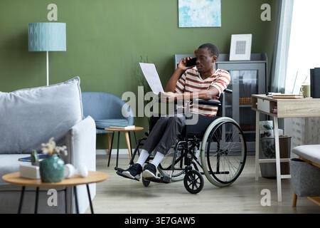 Disabled Black man in wheelchair having phone call and reviewing document report working from home Stock Photo