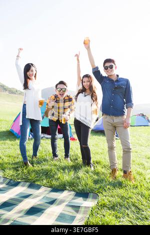 Smiling Chinese male and female friends raising arms and making hand gestures having fun while standing with cups of beer on concert ground at musi... Stock Photo