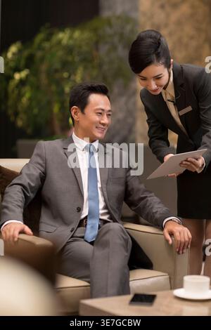Chinese female staff showing menu to Chinese businessman while sitting on sofa in hotel lobby Stock Photo