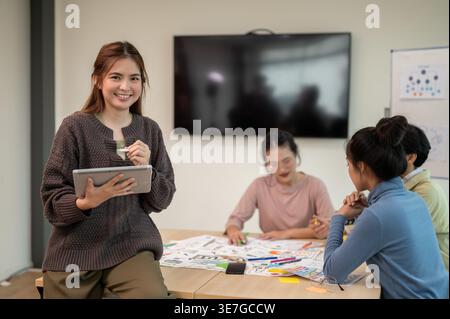 Smiling Asian woman holding tablet sitting on table near workmates in office Stock Photo