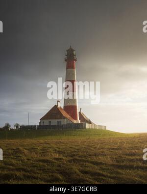 Striped lighthouse in storm light on a grass-covered landscape with threatening clouds, A red and white lighthouse stands on a grassy hill under a clo Stock Photo