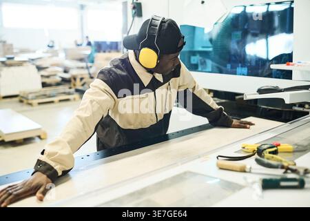 Middle-aged Black male carpenter in protective headphones sawing wooden plank on special machine in factory Stock Photo
