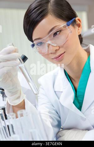 Smiling Asian female doctor wearing protective glasses looking at test tube in laboratory Stock Photo