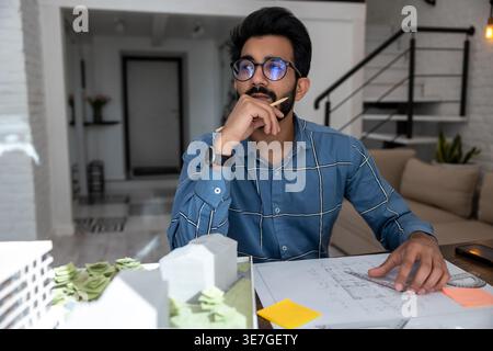 Bearded Indian male architect with eyeglasses leaning chin on hand looking away while drawing plan for building project sitting at drawing table at... Stock Photo