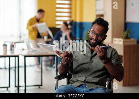 Disabled Asian man holding tablet recording voice message using smartphone sitting in wheelchair in office during daytime Stock Photo
