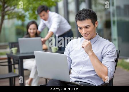 Chinese businessman using laptop sitting at table near colleagues in outdoor cafe Stock Photo