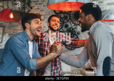 Cheerful Black man having fun and doing manly handshake with caucasian friends standing in bar Stock Photo