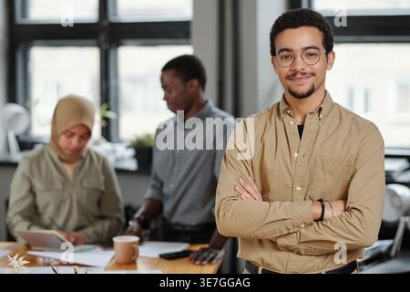 Smiling mixed-race businessman in casualwear standing with crossed arms near male and female colleagues working together in office Stock Photo
