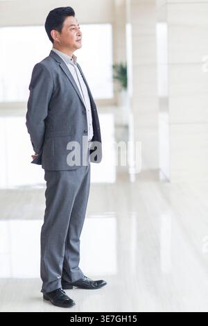 Chinese businessman wearing gray suit looking away while standing with hands behind back near window in office Stock Photo