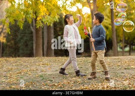 Smiling Chinese boy and girl playing bubbles while standing in autumn park during daytime Stock Photo