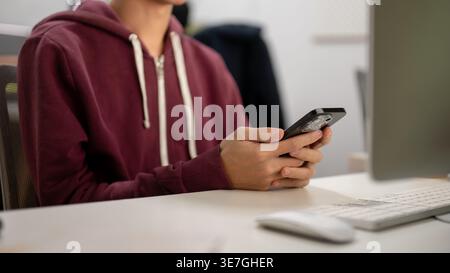 Man wearing maroon hoodie using smartphone while sitting at table in office Stock Photo