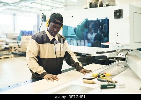 Middle-aged Black male carpenter in protective headphones sawing wooden plank on special machine in factory Stock Photo