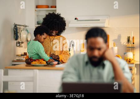 Smiling Black mother with afro standing at kitchen counter playing with her son while husband is working remotely at home Stock Photo