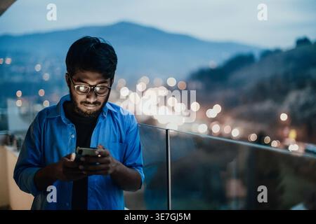 Indian man with beard wearing eyeglasses using smartphone while standing on balcony against city at night Stock Photo