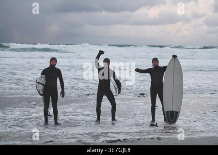 Cheerful Mixed race men wearing surfing suits holding surfboards while standing on snowy beach during winter Stock Photo