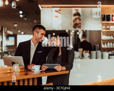 Smiling Hispanic business woman talking to caucasian businessman while using tablet and standing in cafe Stock Photo