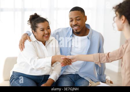 Smiling Black and Hispanic couple shaking hands with female psychologist while sitting on sofa in clinic Stock Photo
