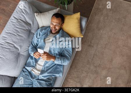 Black man wearing denim jacket using smartphone lying on sofa in living room at home Stock Photo