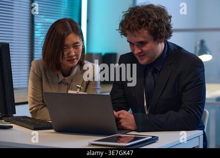 Asian female and caucasian male programmers sitting at desk discussing computer program on laptop working late at night in office Stock Photo