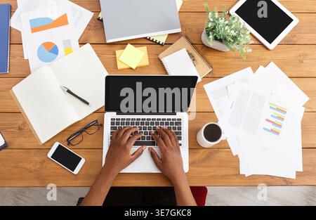 Black businessman working on laptop sitting at wooden desk with various objects in office Stock Photo