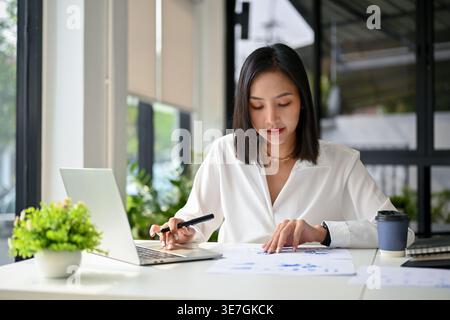 Asian businesswoman looking at documents while working on laptop sitting at table in office Stock Photo